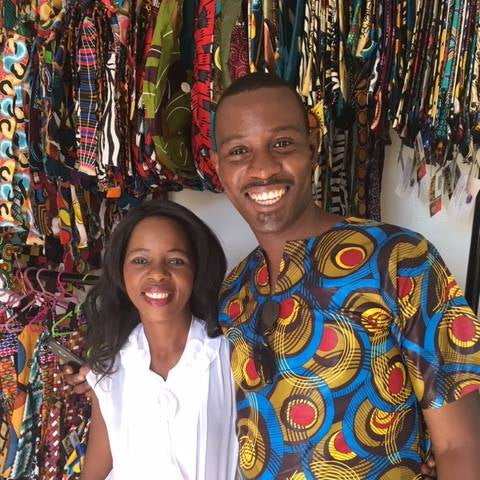Two people wearing colorful traditional attire standing in front of a display of fabric.